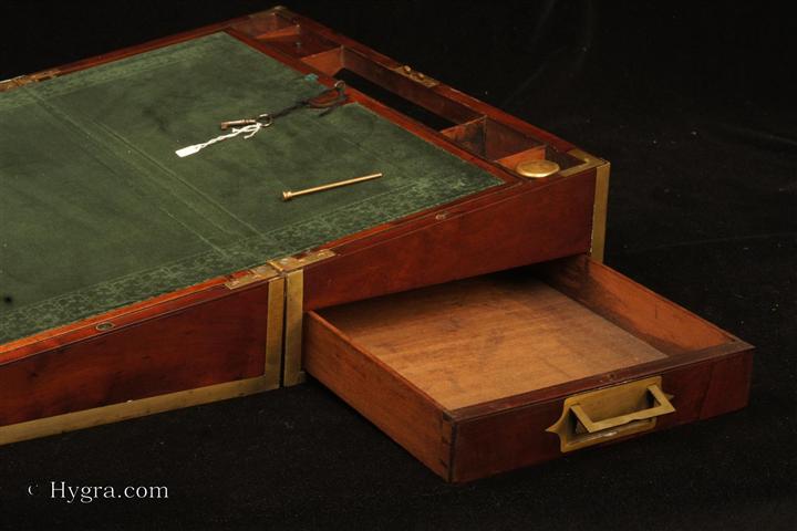 Brass bound solid mahogany writing box of dovetail construction in the military style with countersunk carrying handles a large side drawer secured by a brass pin. The interior has an embossed velvet writing surface  with places for storing papers beneath the flaps (one of which has its own lock). There are also compartments for writing accessories: pens, inkwells. Circa 1815. Enlarge Picture